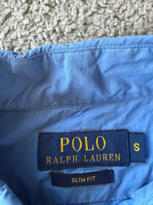 Blå skjorta från Polo Ralph Lauren - Snygg blå skjorta från Polo Ralph Lauren i slim fit-modell. Skjortan har klassisk design med broderad rosa logga på bröstet. Perfekt för dig som gillar stilrena och tidlösa plagg.