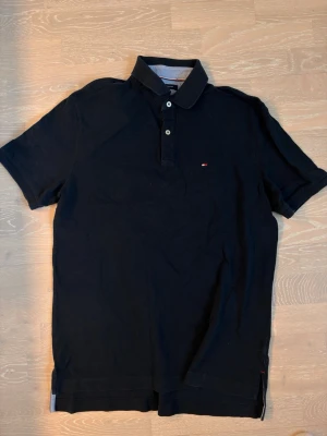 Svart pikétröja från Tommy Hilfiger - Klassisk svart pikétröja från Tommy Hilfiger med korta ärmar och liten logga på bröstet. Tröjan har krage och knappar framtill. Perfekt till jeans eller shorts för en clean och enkel stil.