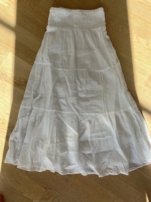 Vit långkjol från Gina Tricot - Luftig och somrig vit långkjol från Gina Tricot med bred resår i midjan och lätt volang nedtill. Kjolen är tillverkad i ett tunt och mjukt bomullstyg som ger en avslappnad och skön känsla. Perfekt för varma dagar och enkel att matcha.