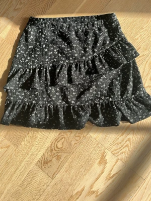 Svart volangkjol med blommigt mönster - Svart kortkjol från Kappahl med små vita blommor och volangdetaljer. Kjolen har resår i midjan och är tillverkad i ett mjukt, lätt material som passar perfekt till en trendig vardagslook.