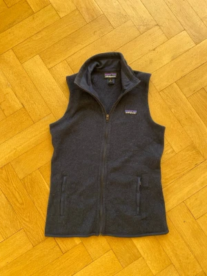 Mörkgrå fleeceväst från Patagonia - Säljer en mörkgrå fleeceväst från Patagonia med hel dragkedja framtill och två fickor med dragkedja. Västen har hög krage och mjukt fleecefoder, perfekt för lager-på-lager. Klassisk Patagonia-logga på bröstet.