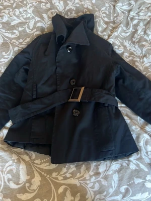 Svart trenchcoat med bälte - Stilren svart trenchcoat med klassisk krage, knäppning framtill och ett matchande tygbälte med fyrkantigt metallspänne. Jackan har två fickor och är tillverkad i ett slätt tyg som ger en snygg siluett. Perfekt för dig som gillar tidlös och enkel stil.