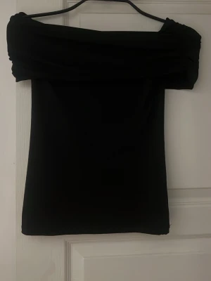 Svart offshoulder topp - Säljer en svart offshoulder topp med bred, vikt kant över axlarna. Toppen är tight och har en stilren, enkel design som passar perfekt till jeans eller kjol. Materialet känns mjukt och stretchigt för en bekväm passform.