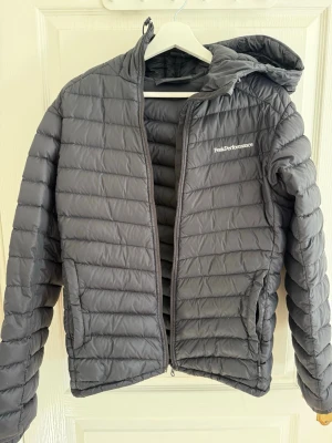 Svart dunjacka från Peak Performance - Snygg svart dunjacka från Peak Performance med huva och dragkedja framtill. Jackan har quiltad design och två sidofickor. Perfekt för kalla vinterdagar och riktigt lätt att bära. Logotypen sitter på bröstet. Priset går diskuteras vid snabbaffär