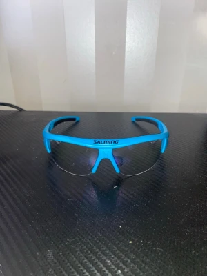 Blå sportiga solglasögon Salming - Säljer ett par Salming V1 Protec Eyewear JR skyddsglasögon i toppskick. Endast använda 3 gånger, så de är i princip som nya!  Perfekta för innebandy och andra sporter där ögonskydd rekommenderas eller är ett krav. Sitter bekvämt, lätt vikt och ger bra skydd utan att påverka sikten.
