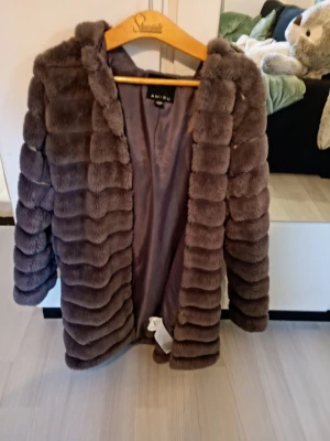 Brun pälsjacka med huva från Amisu - Säljer en brun lång pälsjacka från Amisu med huva och mjukt foder. Jackan har ett fluffigt, ribbat mönster och är perfekt för kalla vinterdagar. Stängs framtill och har en avslappnad passform. Jackan är gjord av syntetisk päls och har en lyxig känsla.