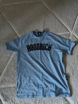Blå t-shirt från Hoodrich - Snygg blå t-shirt från Hoodrich med stor svart logga framtill och coolt blått tryckdetalj över texten. Klassisk passform med korta ärmar och rund hals. Perfekt för en avslappnad streetwear-look.