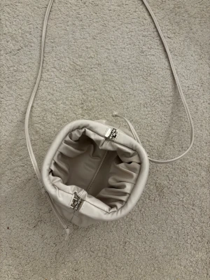 Ljusbeige kuvertväska med dragsko - Trendig ljusbeige kuvertväska med dragsko och lång axelrem. Väskan har en mjuk, rundad form och är tillverkad i ett slätt material som liknar skinn. Perfekt för att bära det viktigaste och ger en stilren look.