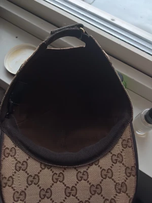 Beige keps från Gucci med GG-mönster - Säljer en beige keps från Gucci med det klassiska GG-monogrammet över hela kepsen. Kepsen har böjd skärm och justerbar rem baktill. Materialet är canvas med detaljer i brunt och inslag av grönt och rött på sidorna. Perfekt för dig som vill ha en lyxig och trendig accessoar.