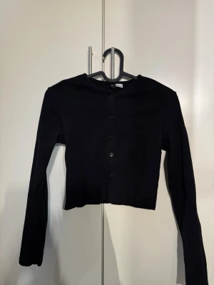 Svart cropped cardigan med knappar - Snygg svart cropped cardigan med långa ärmar och rund halsringning. Tröjan har knäppning framtill med svarta knappar och är tillverkad i ett mjukt, stretchigt material. Perfekt att styla över ett linne eller t-shirt.