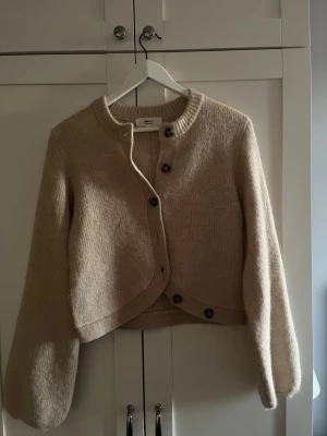 Beige stickad kofta med knappar - Mysig beige stickad kofta med rund halsringning och stora knappar framtill. Koftan har en något kortare, boxig passform och långa, lite puffiga ärmar. Har aldrig använt, bara testad. 