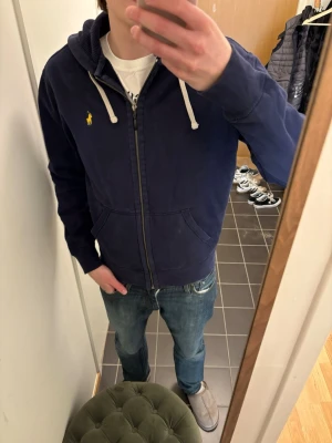 Marinblå hoodie från Polo Ralph Lauren - Snygg marinblå hoodie med dragkedja från Polo Ralph Lauren. Klassisk gul logga på bröstet, vita dragsnören i huvan och två fickor framtill. Tillverkad i mjuk bomullsblandning, perfekt för chill dagar eller när du vill ha en clean look.