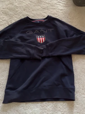 Mörkblå sweatshirt från GANT - Mörkblå sweatshirt från GANT med klassisk logga och sköld med stjärnor och ränder på bröstet. Tröjan har rund halsringning och ribbade muddar. Perfekt för en avslappnad och stilren look.