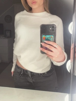 Vit stickad tröja med rund hals - En enkel och stilren vit stickad tröja med rund halsringning och långa ärmar. Tröjan har ribbade muddar vid ärmslut och nederkant. Perfekt att matcha med jeans eller kjol för en avslappnad look.