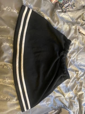 Svart kortkjol med vita ränder - Sportig svart kortkjol från Red & Blue med två vita ränder längs sidan. Kjolen har resår i midjan och är gjord i ett mesh-liknande syntetmaterial som ger en luftig känsla. Perfekt för en avslappnad och trendig look.