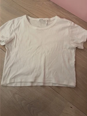 Vit croppad t-shirt från Monki - En basic vit croppad t-shirt från Monki med rund halsringning och korta ärmar. Tillverkad i mjuk bomull, perfekt att styla med jeans eller kjol för en enkel och fräsch look.