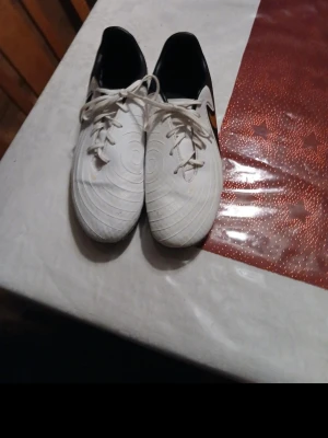 Nike Phantom vita fotbollsskor - Säljer ett par vita Nike Phantom fotbollsskor med svarta detaljer och orange swoosh på sidan. Skorna har klassisk snörning och mönstrad ovansida för bättre bollkontroll. Yttersulan har dobbar som passar för spel på gräs. Perfekta för dig som vill ha stil och funktion på planen.