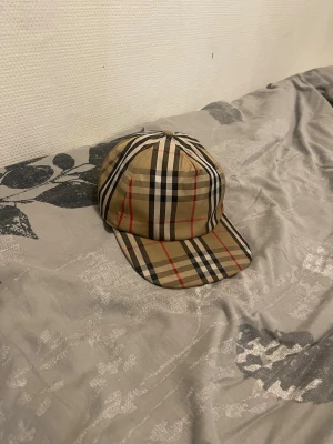 Burberry keps - Säljer en klassisk beige keps från Burberry med det ikoniska rutmönstret i svart, vitt och rött. Kepsen har justerbar snapback och en svart patch med texten 'BURBERRY LONDON ENGLAND' bak. Tillverkad i bomull för en skön och stilren look.