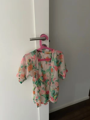 Blommig transparent blus med kort ärm - Söt blus i transparent material med blommigt mönster i rosa, grönt och orange. Modellen har korta ärmar och en luftig passform, perfekt för varma dagar. Blusen har en öppen front och är lätt att matcha med jeans eller kjol.