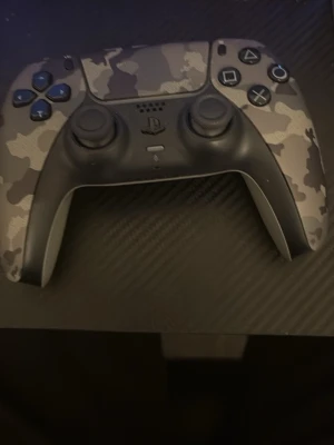 PS5 DualSense Camo handkontroll - Säljer en Sony PS5 DualSense handkontroll med grått camo-mönster. Kontrollern har klassisk PlayStation-design med två analoga spakar, styrkors och touchpad. Tillverkad i plast med matta och blanka detaljer. Perfekt för gaming på PlayStation 5.