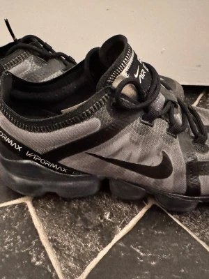 Nike Air Vapormax svarta sneakers - Säljer ett par Nike Air Vapormax sneakers i svart och grått med tydlig logga på sidan och textdetaljer på plösen. Skorna har en genomskinlig sula och ovandel i mesh-liknande material med svarta snören. Snygg och sportig design som sticker ut.