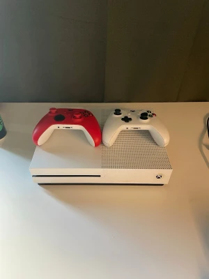 Xbox One S med två handkontroller - Säljer en vit Xbox One S med två trådlösa handkontroller, en röd och en vit. Konsolen är fullt fungerande. Ni får med en massa spel också så som FIFA, gta mm