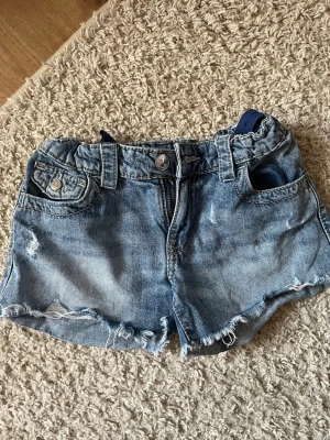 Blå denimshorts med slitningar - Säljer ett par blå denimshorts med råa kanter och slitningar. Shortsen har klassiska fickor fram och bak, knappstängning och bälteshällor. Perfekt för en avslappnad och cool stil under varma dagar.