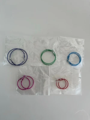 5 par Färgglada örhängen  - Säljer fem par tunna hoop örhängen i olika färger: blå, grön, ljusblå, rosa och röd. Alla örhängen är runda och tillverkade i metall med enkel låsmekanism. Perfekta för att addera färg till din stil!