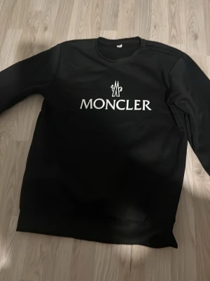 Svart Moncler sweatshirt - Svart sweatshirt från Moncler med vit logotyptryck på bröstet. Klassisk rund halsringning och långa ärmar. Perfekt för en stilren och trendig look.