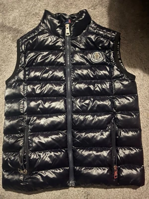 Moncler väst - Snygg svart dunväst från Moncler med glansig finish och quiltad design. Västen har två dragkedjeförsedda fickor, hög krage och Moncler-logga på bröstet. Perfekt för lager på lager och streetstyle. Kan gå ner i pris och ställ frågor om ni undrar något😉