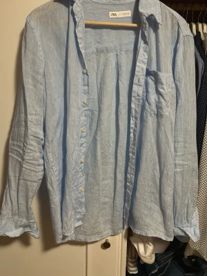 Ljusblå linneskjorta från Zara - Snygg ljusblå skjorta från Zara i luftigt linne. Skjortan har klassisk krage, bröstficka och knappar framtill. Perfekt för en avslappnad och stilren look. Långärmad modell med normal passform.