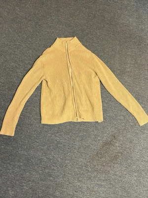 Beige ribbad ziptröja - Snygg beige ribbstickad tröja med dragkedja framtill. Tröjan har långa ärmar och är tillverkad i ett mjukt, stretchigt material som sitter skönt på kroppen. Perfekt för lager-på-lager och enkel att matcha med jeans.