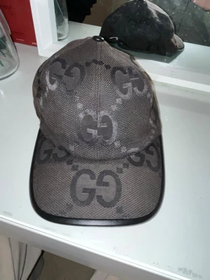 Gucci keps med GG-mönster - Snygg keps från Gucci i mörkbrunt tyg med det klassiska GG-monogrammet över hela kepsen. Kepsen har böjd skärm med svart kant och justerbar passform. Tillverkad i Italien och perfekt för dig som gillar exklusiva accessoarer.