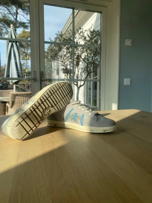 Premiata Skor - Snygga gråa sneakers från Premiata med blå detaljer på sidan. Skorna har en klassisk låg siluett, vita snören och en beige sula. Ovandelen är i mocka och textil för en stilren look.