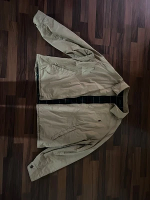 Beige vintage vindjacka från Polo Ralph Lauren - Stilren beige vindjacka från Polo Ralph Lauren med klassisk krage och dragkedja framtill. Jackan har två snedställda fickor och diskret broderad logga på bröstet. Insidan har rutigt foder för extra stilpoäng. Perfekt för svala dagar. Jackan har också en till ficka på insidan, så totalt 3 fickor. Storlek är 2XB och passar som loose XL. Skriv om du har några frågor!