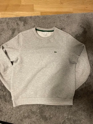 Grå sweatshirt från Lacoste - Snygg grå sweatshirt från Lacoste med klassisk rund hals och det ikoniska gröna krokodilmärket broderat på bröstet. Tröjan har ribbade muddar vid ärmslut och nederkant. Perfekt för en avslappnad och stilren look.