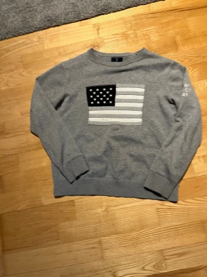 Grå stickad gant tröja från Gant - Snygg grå stickad tröja från Gant med amerikansk flagga i vitt och grått på bröstet. På ärmen finns brodery med texten 'GANT NH-CT 194 playground'. Tröjan har rund hals och ribbade muddar vid ärmslut och nederkant. Perfekt för en avslappnad stil.
