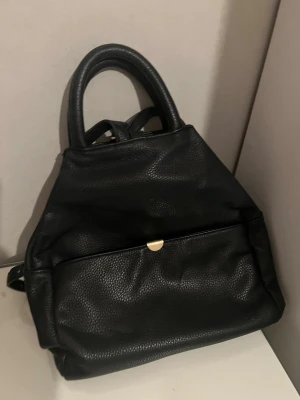 Svart ryggsäck i skinnimitation - Snygg svart ryggsäck i skinnimitation med guldfärgade detaljer. Väskan har ett stilrent, trekantigt snitt, dubbla handtag och justerbara axelremmar. Framtill finns en ficka med tryckknapp och huvudfacket stängs med dragkedja. Perfekt för dig som vill ha en trendig och praktisk väska.