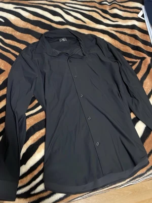 Svart skjorta från H&M - Stilren svart skjorta från H&M med klassisk krage och knäppning framtill. Skjortan har lång ärm och är tillverkad i ett mjukt och lätt material som passar perfekt till olika outfits.