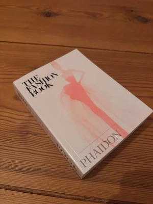 The Fashion Book av Phaidon - En snygg och inspirerande bok om mode, 'The Fashion Book' från Phaidon. Omslaget är vitt med en stiliserad illustration i rosa och svart text. Perfekt för dig som älskar mode och vill ha en cool coffee table-bok. Den är pytteliten - max 20 cm i höjd.