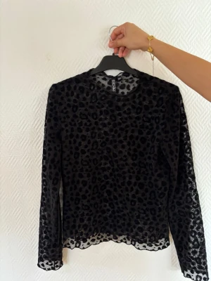 Svart mesh topp med leopardmönster - Snygg svart mesh topp från Pieces med transparent tyg och flockat leopardmönster. Långärmad modell med rund halsringning, perfekt för att skapa en trendig och edgy look. Tunn och lätt, passar bra att styla i lager.