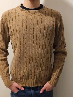 Beige kabelstickad tröja Gant - Snygg beige kabelstickad tröja från Gant med rund halsringning och diskret logga broderad på bröstet. Tröjan har ribbade muddar vid ärmslut och nederkant, perfekt för lager på lager och en klassisk stil.