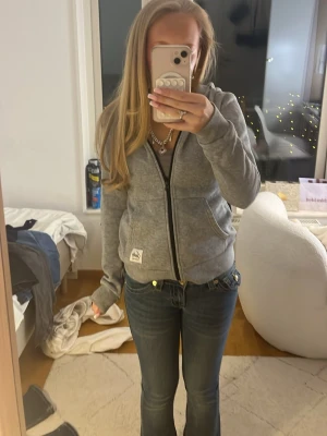 Grå zip-hoodie från Andy - Säljer en grå hoodie från Hollister med dragkedja och huva. Hoodien har ribbade muddar, två fickor framtill och ett litet Hollister-märke på nedre kanten. Perfekt för dig som gillar en avslappnad och stilren look.