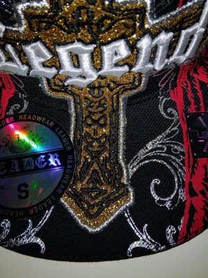 Svart keps med brodyr från Leader - Säljer en svart keps från Leader med stor broderad text och kors i guld, silver och vitt framtill. Kepsen har även röda detaljer och coola mönster på sidorna. Märkesskylt och originaletikett sitter kvar. Perfekt för dig som gillar streetwear och unika accessoarer.