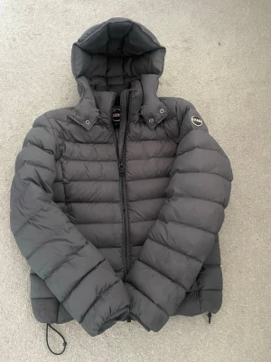 Colmar jacka  - Snygg svart dunjacka från Colmar med huva och dragkedja framtill. Jackan har två fickor med dragkedja och Colmar-logga på ena ärmen. Perfekt för kalla vinterdagar tack vare det vadderade materialet.