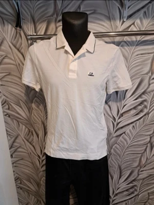 Vit piké från C.P. Company - Klassisk vit pikétröja från C.P. Company med korta ärmar och krage. Tröjan har knappslå framtill och en liten logotyp på bröstet. Perfekt till jeans eller shorts för en clean och enkel look.