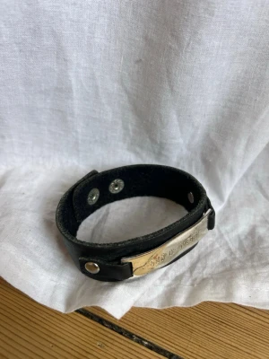 Svart läderarmband med metallplatta - Coolt svart armband i läder med en silverfärgad metallplatta som har ingraverad text. Armbandet har tryckknappar för enkel justering och en stilren design som passar till många outfits.