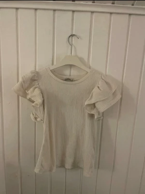 Vit ribbad topp med volangärmar - Supersöt vit ribbad topp med stora volangärmar som ger en extra touch till outfiten. Rund halsringning och figurnära passform. Perfekt att styla med jeans eller kjol för en trendig look.