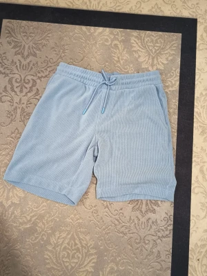 Ljusblå frottéshorts med snörning - Snygga ljusblå shorts i mjuk frotté med elastisk midja och snörning framtill. Shortsen har sidofickor och en bakficka, perfekt för en chill sommarlook. Avslappnad passform och sköna att bära hela dagen.