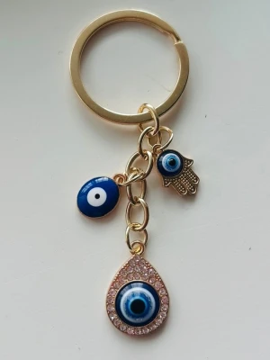 Nyckelring med blått öga och hamsa - Snygg nyckelring i guldfärgad metall med blått öga och hamsa-hand som hängen. Dekorerad med gnistrande stenar runt det stora ögat och detaljer i blått och vitt. Perfekt accessoar för att piffa upp väskan eller nyckelknippan.